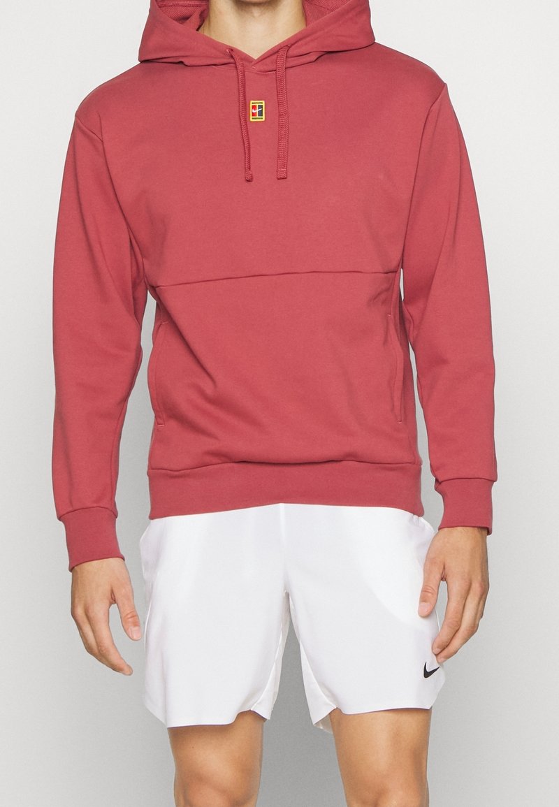 Homme portant un sweat à capuche rouge avec cordons et un short blanc de sport avec un petit logo Nike noir sur la jambe gauche.