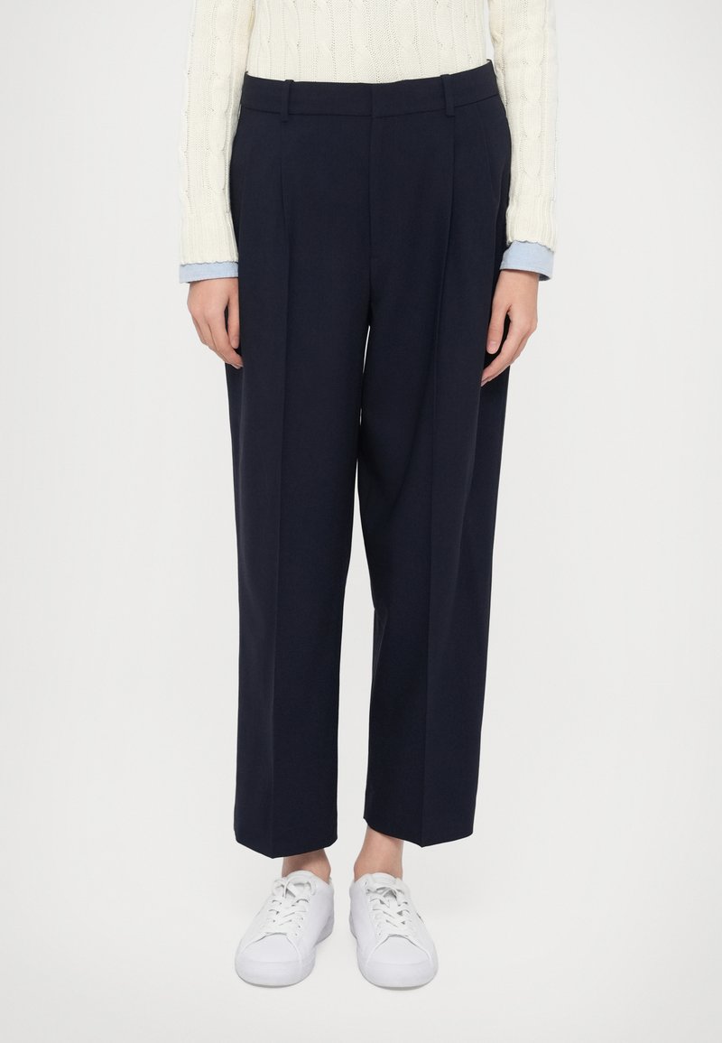 Temno modre široke hlače z naborki spredaj, krojene po meri, združene z belimi supergami. Tekstura je videti gladka, tkanina pa ima strukturiran videz.