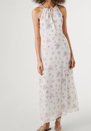 Kobieta ubrana w białą sukienkę maxi na szyję z małymi lawendowymi kwiatowymi nadrukami i beżowe płaskie sandały na paski.
