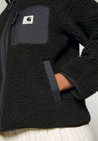 Czarna polarowa kurtka o teksturowej powierzchni, z zamkiem z przodu, bocznymi kieszeniami oraz kontrastową nylonową kieszenią na klatce piersiowej z logo.