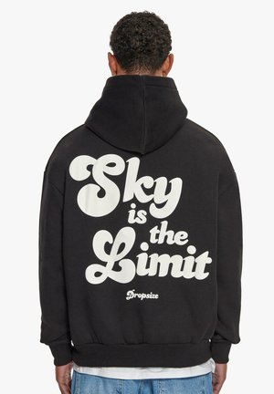 Person med kort krøllet hår iført sort hættetrøje med teksten "Sky is the Limit" skrevet med store hvide bogstaver på ryggen.