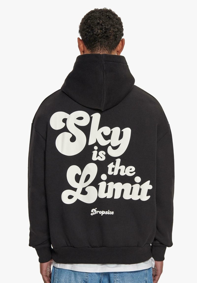 Personne aux cheveux courts et bouclés portant un sweat à capuche noir avec l'inscription "Sky is the Limit" en grandes lettres blanches dans le dos.