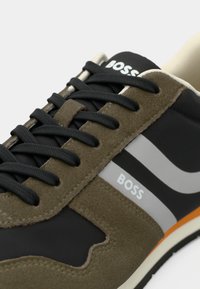 Grüner und schwarzer Sneaker aus Wildleder und synthetischen Materialien, ausgestattet mit schwarzen Schnürsenkeln, silbernen Akzenten und einer auffälligen orangen Sohle. "BOSS"-Logo sichtbar.