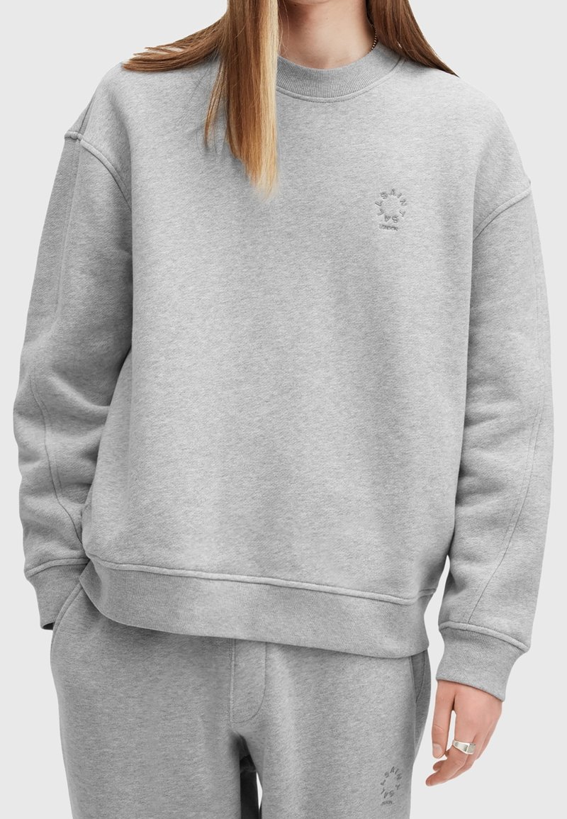 Personne portant un sweat-shirt gris clair et un pantalon de survêtement assorti avec un logo circulaire subtil sur la poitrine et la cuisse, la main reposant le long du corps.