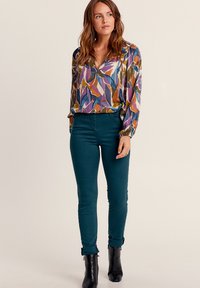 Blusa multicolore con motivo a foglie, scollatura a V e maniche lunghe, abbinata a jeans skinny turchesi e stivaletti neri.