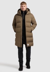 Karamelkrāsas puffer jaka ar kapuci, ar kviltētu pildījumu un priekšējo rāvējslēdzēju. Valkāta virs melna turtlenecka un kopā ar melniem biksēm un zābakiem.