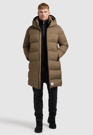 Teddyjack in tan met een capuchon, voorzien van een gewatteerde voering en een ritssluiting aan de voorkant. Draagt over een zwarte coltrui en gecombineerd met zwarte broek en laarzen.