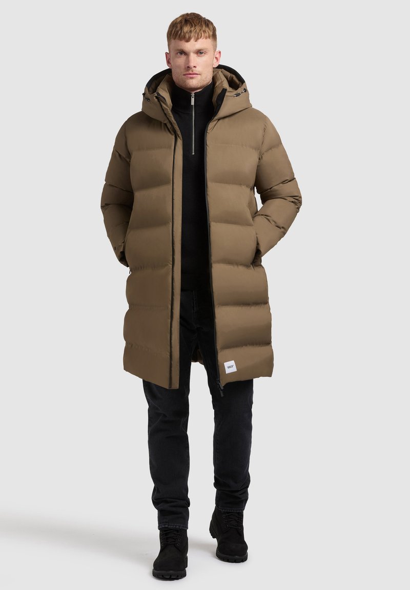 Karamelkrāsas puffer jaka ar kapuci, ar kviltētu pildījumu un priekšējo rāvējslēdzēju. Valkāta virs melna turtlenecka un kopā ar melniem biksēm un zābakiem.