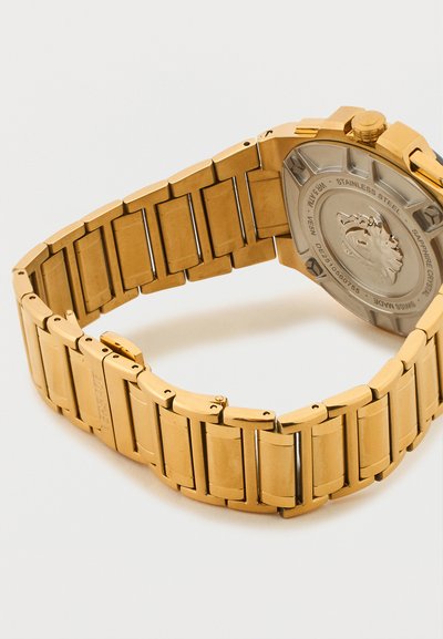 Aukso spalvos metalinė laikrodžio apyrankė su sagtimi, rodanti nerūdijančio plieno nugarėlę, išgraviruotą liūto emblema ir tekstu.