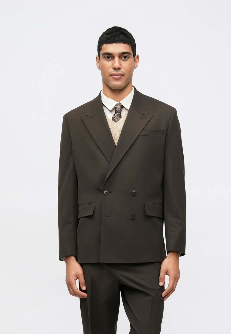 Brown double-breasted suit jacket with notched lapels, two front pockets, worn over a cream knitted vest, paired with a tie and shirt.