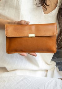 Woman in white dress holding a slim tan leather clutch purse with gold clasp at chest level.