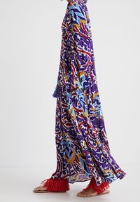 Robe longue colorée avec une base violette, des motifs floraux et géométriques en rouge, bleu et jaune, manches à volants et sandales rouges à franges.