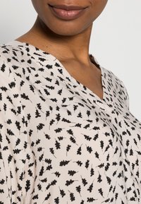 Hellbeige Bluse mit V-Ausschnitt, die ein schwarzes Blattmuster zeigt. Weicher Stoff mit glatter Textur und einer lockeren Passform am Halsauschnitt.