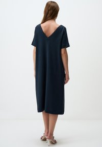 Marineblaues Leinenkleid mit V-Ausschnitt, kurzen Ärmeln und lockerer Silhouette. Glatte Textur, knielang mit seitlichen Schlitzen. Silberne Absatzsandalen.