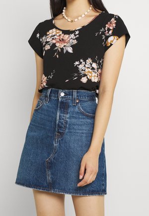 Woman wearing black floral short-sleeve blouse tucked into high-waisted blue denim skirt with raw hem, and shell necklace.