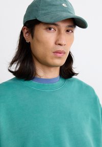 Grønne kordfløjlskasket med et logo, båret med en teal sweatshirt med en blå rund hals. Glat tekstur og afslappet pasform.