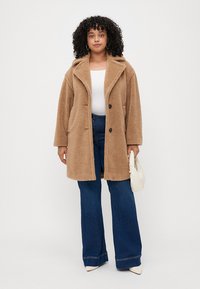 Casaco teddy castanho, com uma textura suave, grande gola e fechos em botão, vestido sobre uma blusa branca e jeans azul de perna larga. Acessório de bolsa branca.