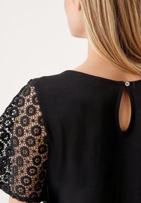 Haut noir avec des manches en dentelle délicate ornées de motifs floraux et un petit détail en forme de trou à l'arrière, fabriqué à partir d'un tissu doux et texturé.