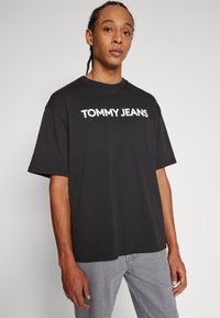Camiseta negra de manga corta con cuello redondo, que presenta el texto en blanco "TOMMY JEANS" en el pecho, confeccionada en tejido de algodón con una textura suave.