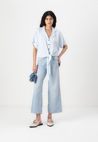 Light blue, short-sleeved striped blouse with a knotted front, paired with wide-leg light denim jeans and beige shoes. Holds a blue accessory.