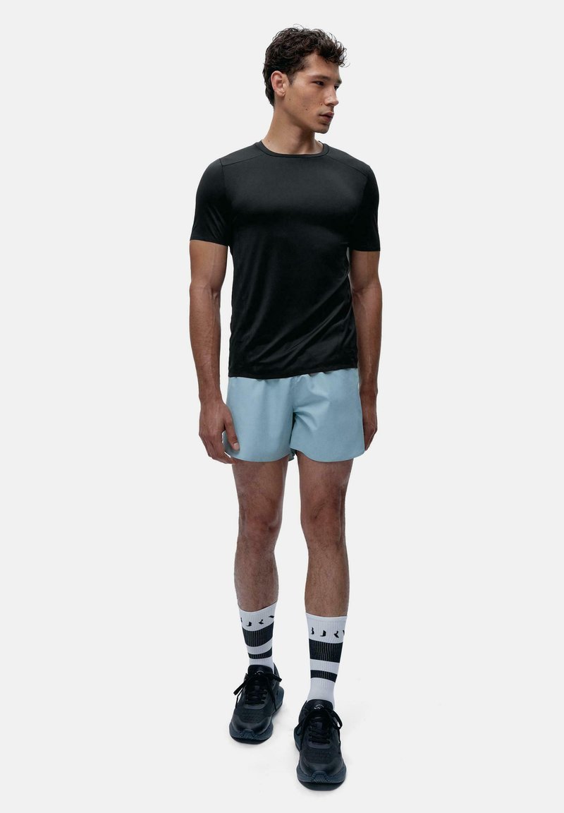 Melnā sporta t-kreklā, gaiši zilajos šortos, melnās sporta apavās un rakstainās zeķēs. Uzsvars uz piemērotību un komfortu minimālisma dizainā.