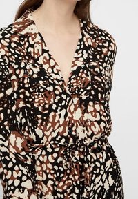 Camicia a fantasia marrone, nera e crema; presenta un colletto, due tasche sul petto e una cintura in vita. Tessuto liscio con un aspetto testurizzato.