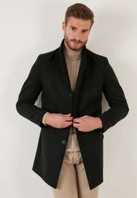 Abrigo de mezcla de lana negro con un corte entallado, cuello con muesca, cierre de un solo botón y un bolsillo en el pecho izquierdo. Llevado sobre un suéter de punto beige.