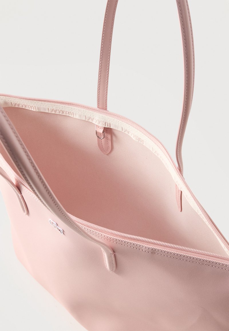 Borsa tote rosa chiaro con manici sottili, apertura superiore che mostra una fodera interna liscia e una cerniera in tessuto lungo il bordo di apertura.