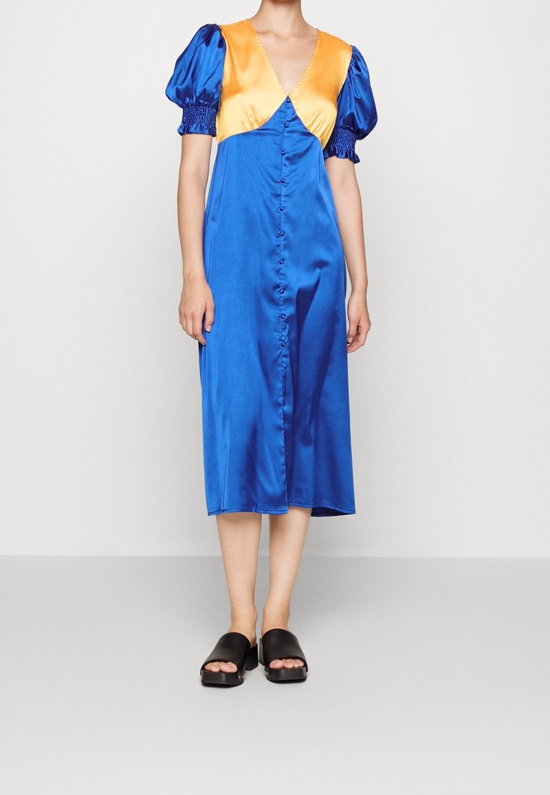 Robe en satin avec un décolleté en V profond, corps bleu et manches bouffantes orange. Elle comporte une fermeture boutonnée à l'avant et arrive au milieu du mollet, associée à des mules noires.
