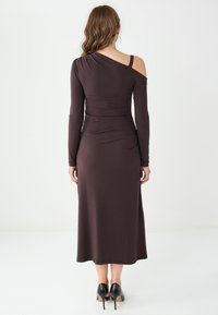 Langes braunes Kleid mit langen Ärmeln und einer freiliegenden Schulter, taillierte Silhouette, weicher Stoff und Midi-Länge, kombiniert mit schwarzen High-Heels.