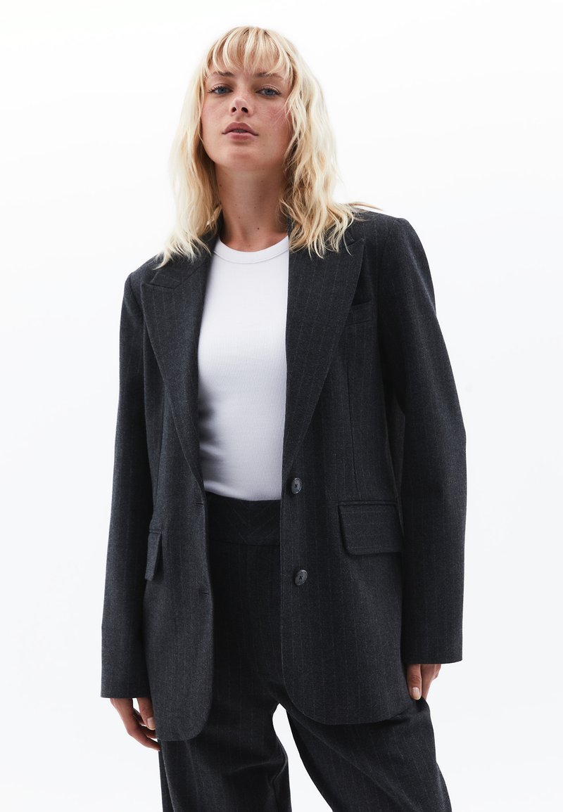 Sort blazer med pinstripes, med reverskrave, to knapper og forlomme, lagd over en hvid tætsiddende skjorte i glat tekstur.