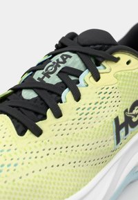 HOKA hardloopschoen met een ademende gele mesh bovenkant, zwarte veters en een gevoerde witte zool. Heeft een textuurpatroon en logodetail.