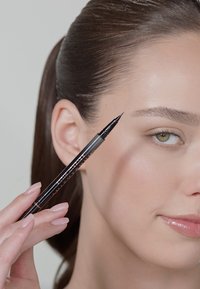 Black brow pencil held by a person with natural nails, placed near the brow area of a light-skinned individual with straight hair.