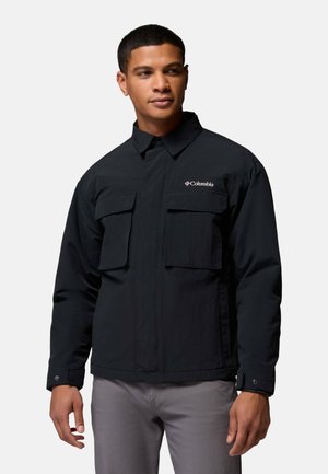 Mann trägt eine schwarze Columbia-Jacke mit zwei Brusttaschen und graue Hose, steht vor einem einfarbigen hellen Hintergrund.
