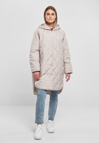 Quiltet lang jakke i lys beige med hette, med knappelukking, sidelommer og en avslappet passform over lyseblå jeans.
