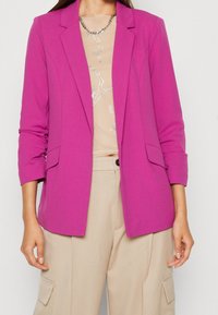 Fuchsia blazer i en slimmad design med slag och två framfickor, buren över en beige topp med ett silvermönster. Mjuk tygtextur.