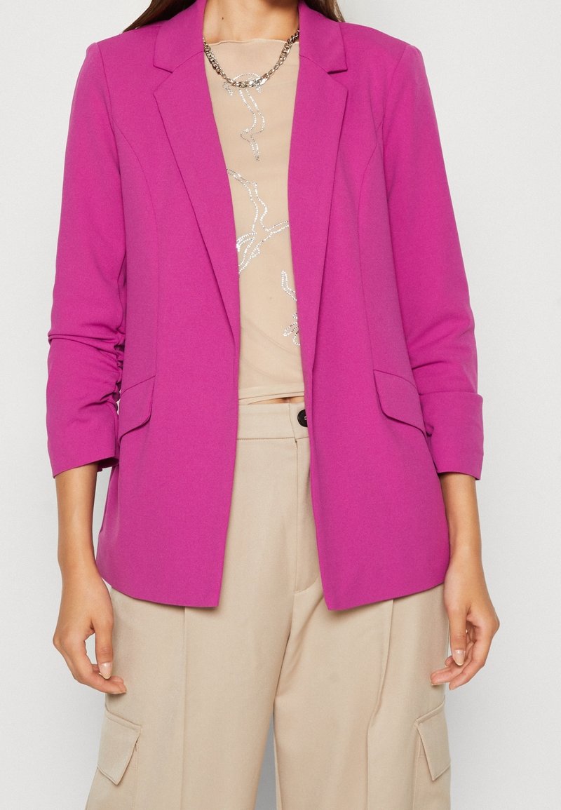 Fuchsia blazer i en slimmad design med slag och två framfickor, buren över en beige topp med ett silvermönster. Mjuk tygtextur.