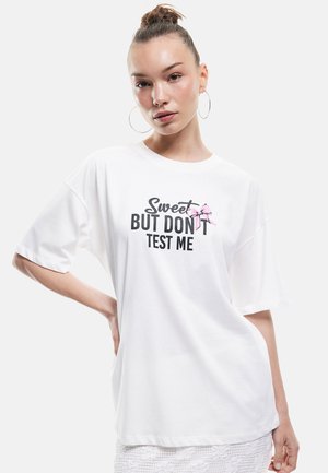 Witte oversized t-shirt met zwarte en roze tekst: "Sweet BUT DON'T TEST ME" naast een roze strik. Zachte katoenen stof, ontspannen pasvorm.