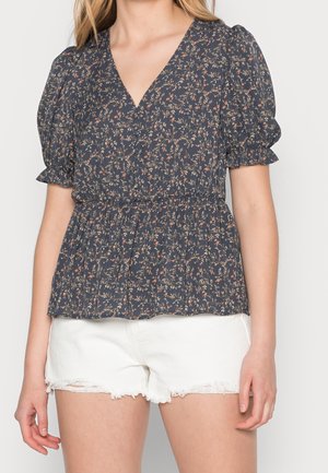 Kobieta w granatowej, kwiatowej bluzce z krótkim rękawem z bufkami oraz białych, postrzępionych szortach jeansowych, stojąca na jednolitym tle.