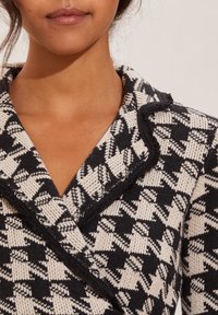 Neulepaita, jossa on musta-valkoinen houndstooth-kuvio. Varustettu kaarrokeella ja repaleisilla reunoilla, mikä korostaa tekstuurista viimeistelyä.