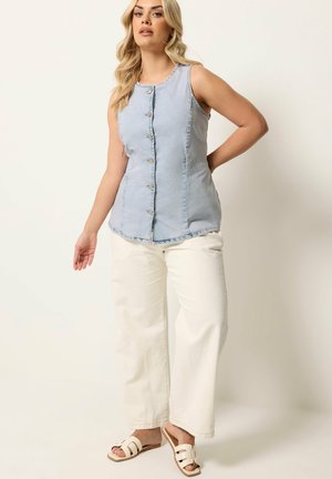 Woman standing wearing sleeveless light blue buttoned top, wide white pants, and white slide sandals against plain background.