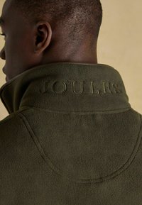 Zielona polarowa kurtka z wysokim kołnierzem i wytłoczonym logo "JOULES". Miękka tekstura i schludne szwy na kołnierzu.