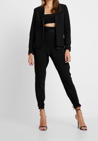Traje negro a medida que incluye una blazer con un solo botón, un top que deja el abdomen descubierto y pantalones entallados con puños ajustables. Completado con tacones altos.