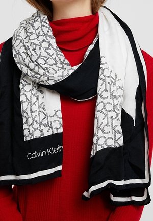 Femme portant une écharpe Calvin Klein noire et blanche avec imprimé logo, sur un pull à col roulé rouge.
