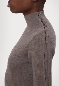 Gros plan d'une personne portant un pull gris côtelé à col haut avec une rangée de boutons le long de l'épaule et de la manche.