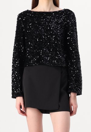 Sort top med lange ærmer med tætte sorte palietter kombineret med en sort asymmetrisk mininederdel på en stående person.