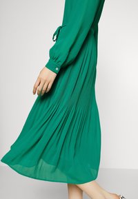 Zelené dlouhé šaty s dlouhým rukávem a zavazováním v pase, s plisovanou texturou u spodního lemu a knoflíkovými manžetami, vyrobené z lehkého materiálu.