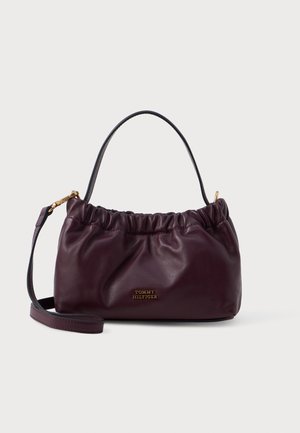 Burgundijas krāsas ādas rokassoma ar sakropļotu dizainu, zelta metāla akcentiem un noņemamu siksnu; priekšpusē ir logotipa detalizācija.