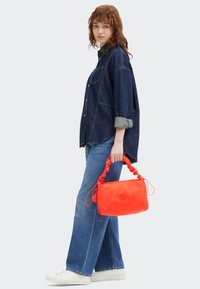 Borsa a tracolla in nylon rosso con manico arricciato, caratterizzata da un logo circolare. Il modello indossa una camicia di denim e jeans a gamba larga, abbinati a sneakers bianche.