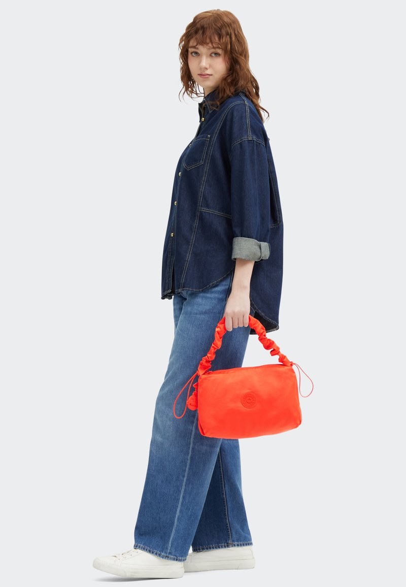 Rode nylontas met een geplooide handgreep, met een circulair logo. Model draagt een denim shirt en wijde jeans, met witte sneakers.